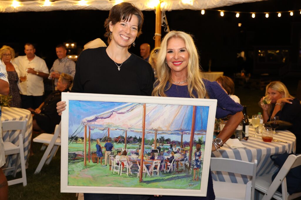 two women holding up a painting at an outdoor event
